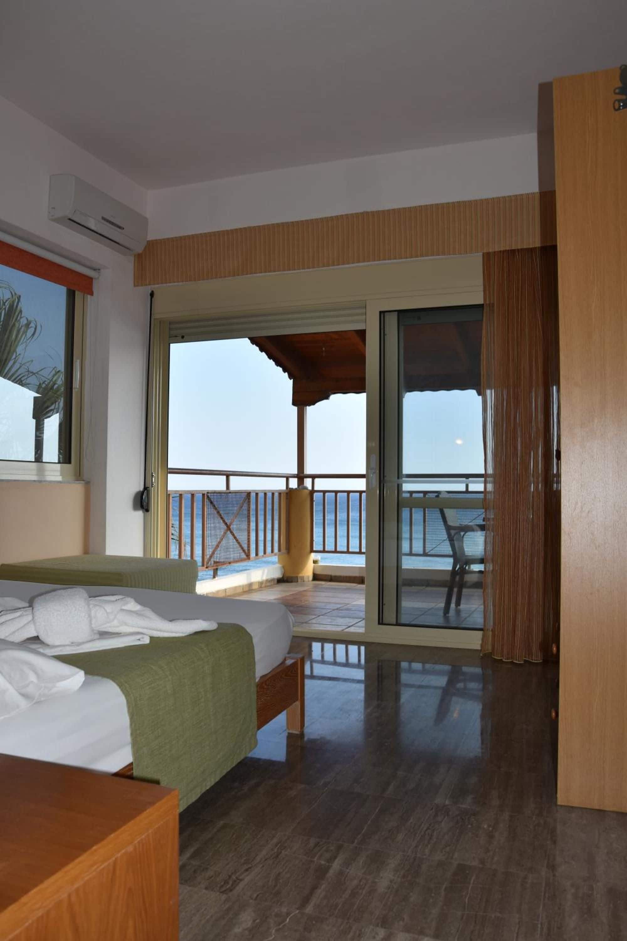 A hotel room with a bed, a wooden desk, a balcony with a view of the sea, and a sliding glass door.