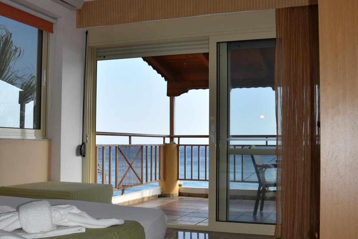 Een hotelkamer met een bed, een houten bureau, een balkon met uitzicht op zee en een glazen schuifdeur.