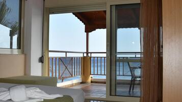 A hotel room with a bed, a wooden desk, a balcony with a view of the sea, and a sliding glass door.