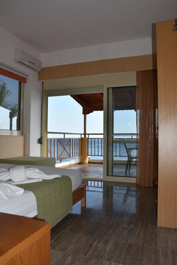 A hotel room with a bed, a wooden desk, a balcony with a view of the sea, and a sliding glass door.