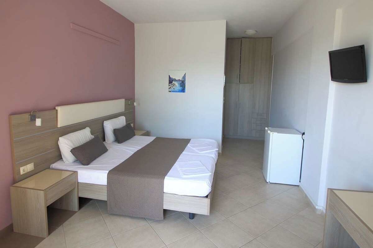 Une chambre à coucher avec un lit, des tables de chevet, un petit réfrigérateur blanc, un téléviseur fixé au mur et un tableau accroché au mur.