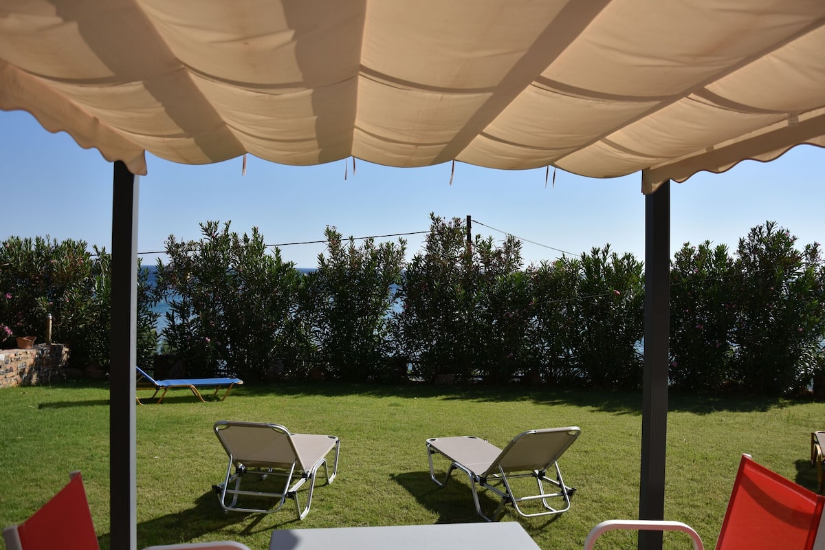 A covered outdoor area with two lounge chairs and a table, surrounded by a hedge and a view of the sea.