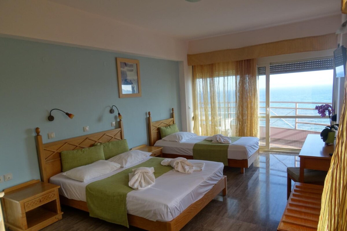Hotelkamer met twee bedden, een houten hoofdbord, een klein tafeltje en uitzicht op zee door grote ramen.