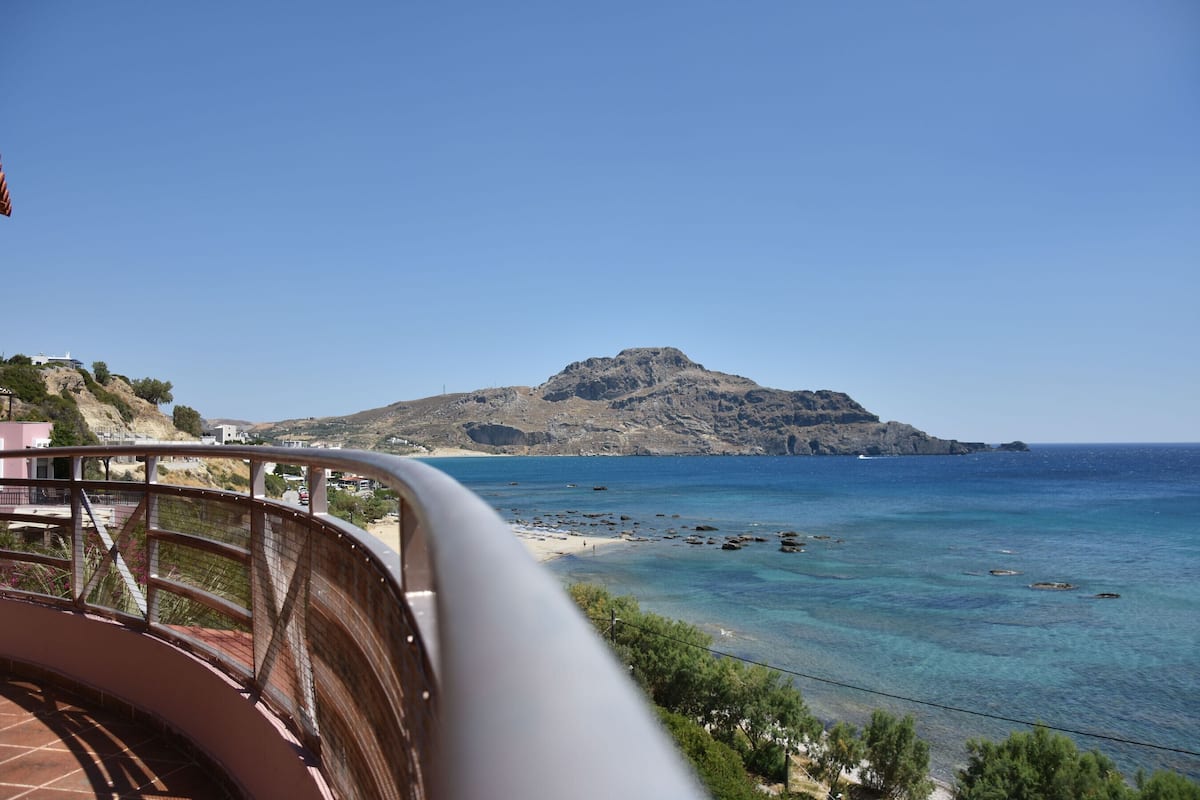 A coastal view with a mountain, clear blue sea, and a balcony with a railing.