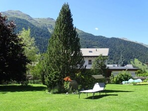 Ein Haus mit Rasen, einem hohen Baum und Bergen im Hintergrund.