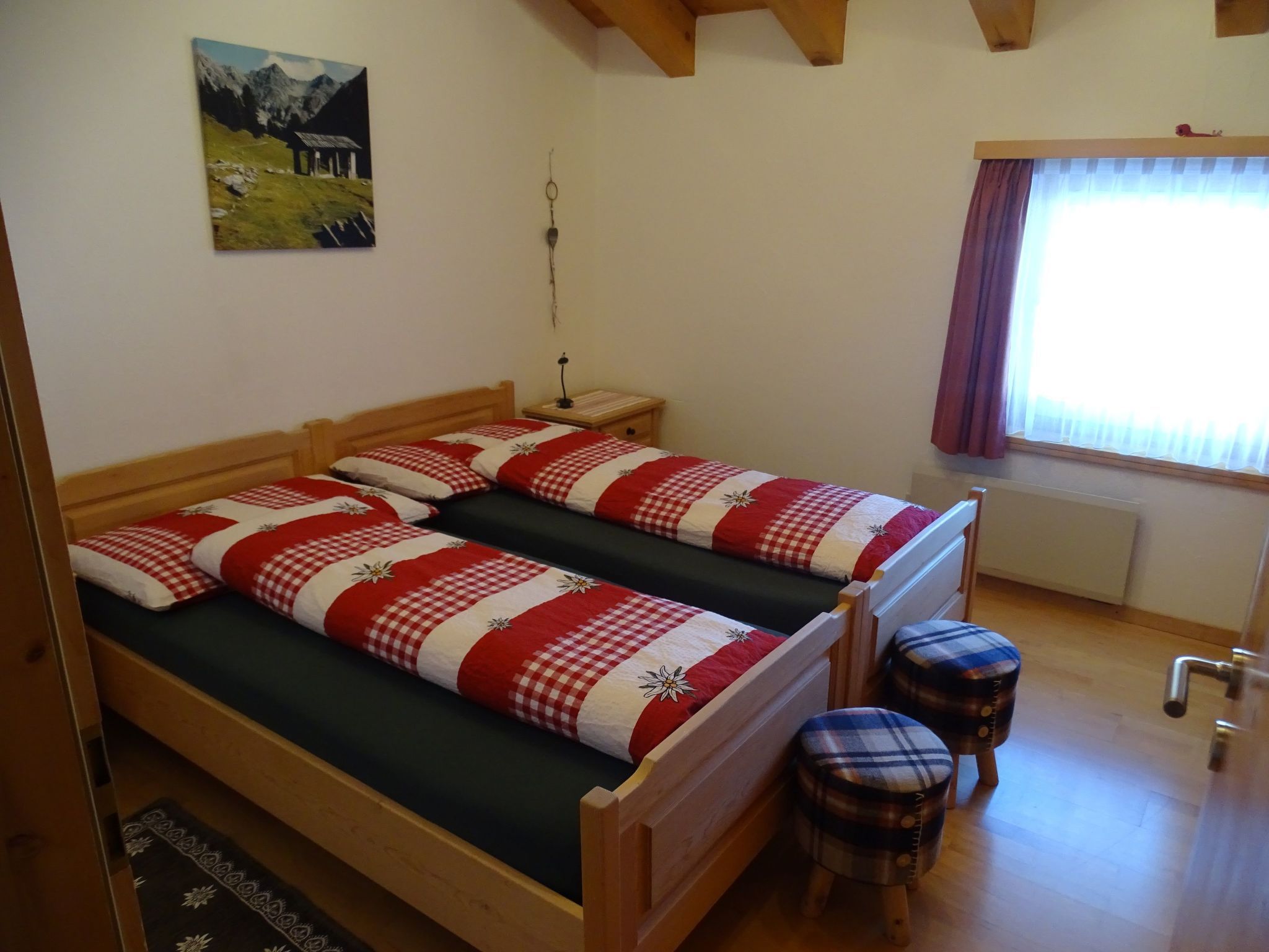Ein Zimmer mit zwei hölzernen Betten, einem kleinen Hocker, einem Nachttisch, einer Lampe und einem fenster mit Vorhängen.