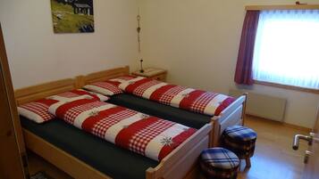 Ein Zimmer mit zwei hölzernen Betten, einem kleinen Hocker, einem Nachttisch, einer Lampe und einem fenster mit Vorhängen.