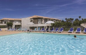 A swimming pool with lounge chairs, a building with a tiled roof, and palm trees in the background. - Residence Odalys Acqua Linda (Poggio-Mezzana)