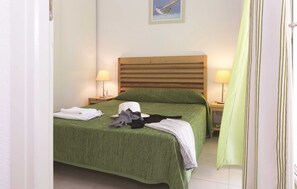 Un lit bien fait, recouvert d’une couverture verte, avec deux lampes de chevet et un tableau encadré accroché au mur.