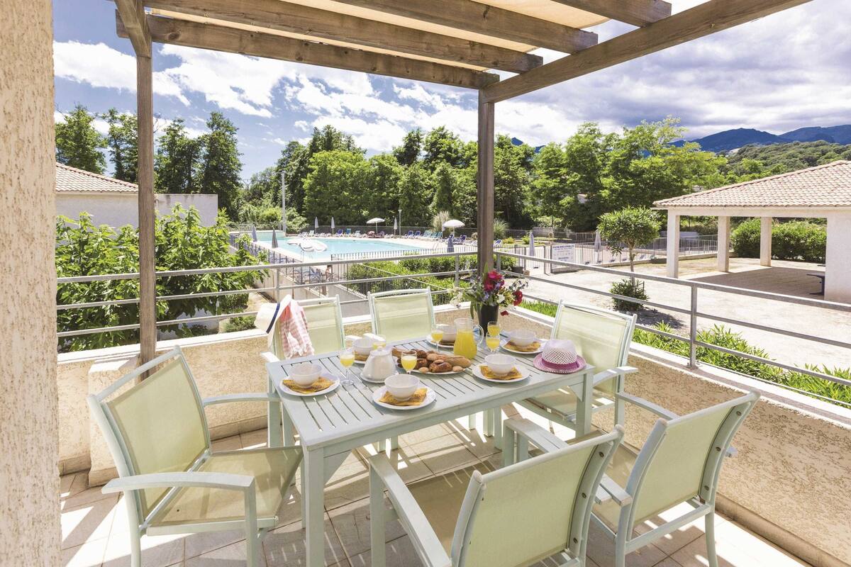Uma área de refeições coberta ao ar livre, vista para piscina, cadeiras brancas e mesa posta para o café da manhã.