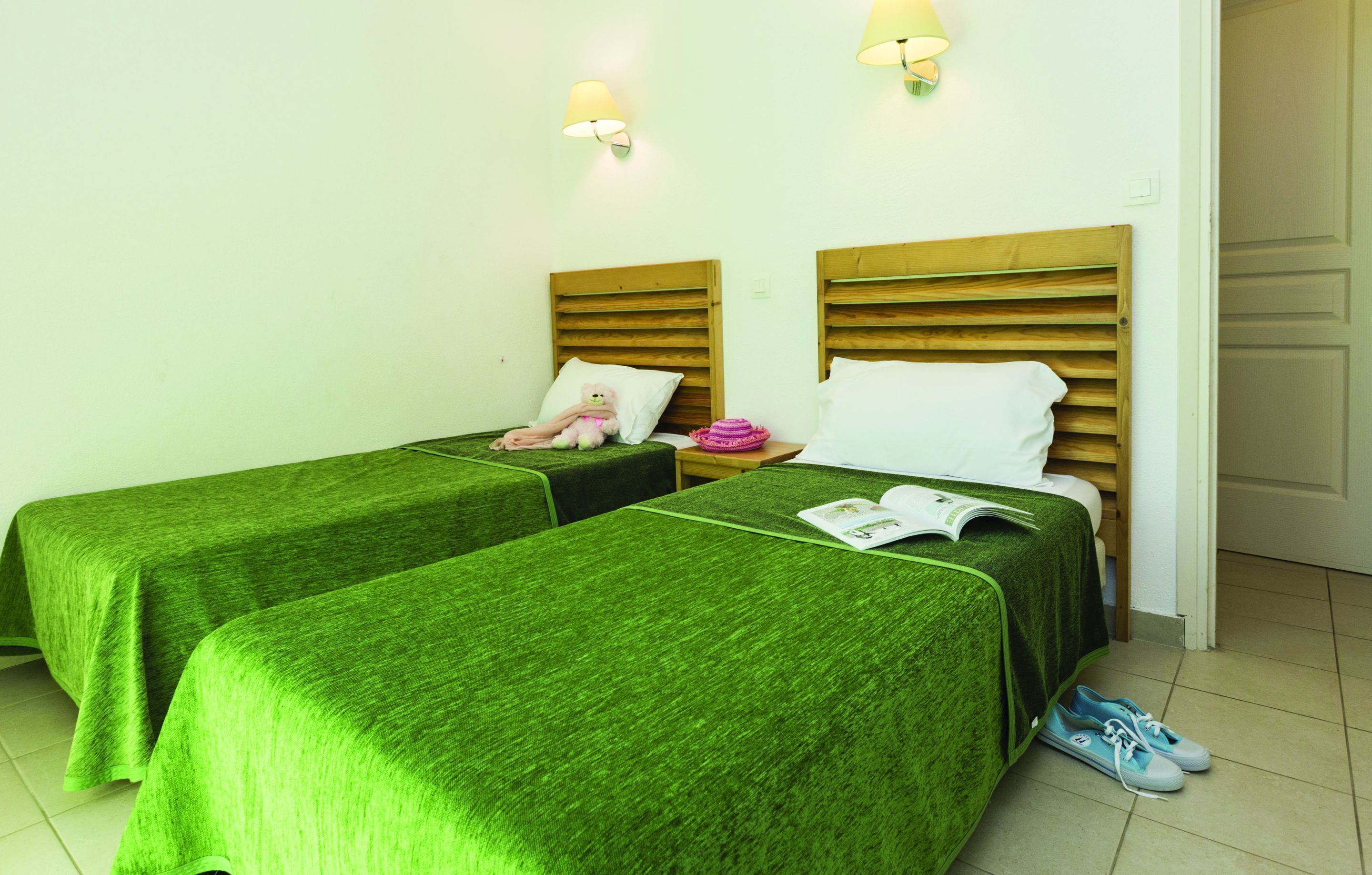 Deux lits simples avec des couvre-lits verts, des têtes de lit en bois, des lampes de chevet, un ours en peluche, un chapeau rose et un magazine sur l’un des lits.