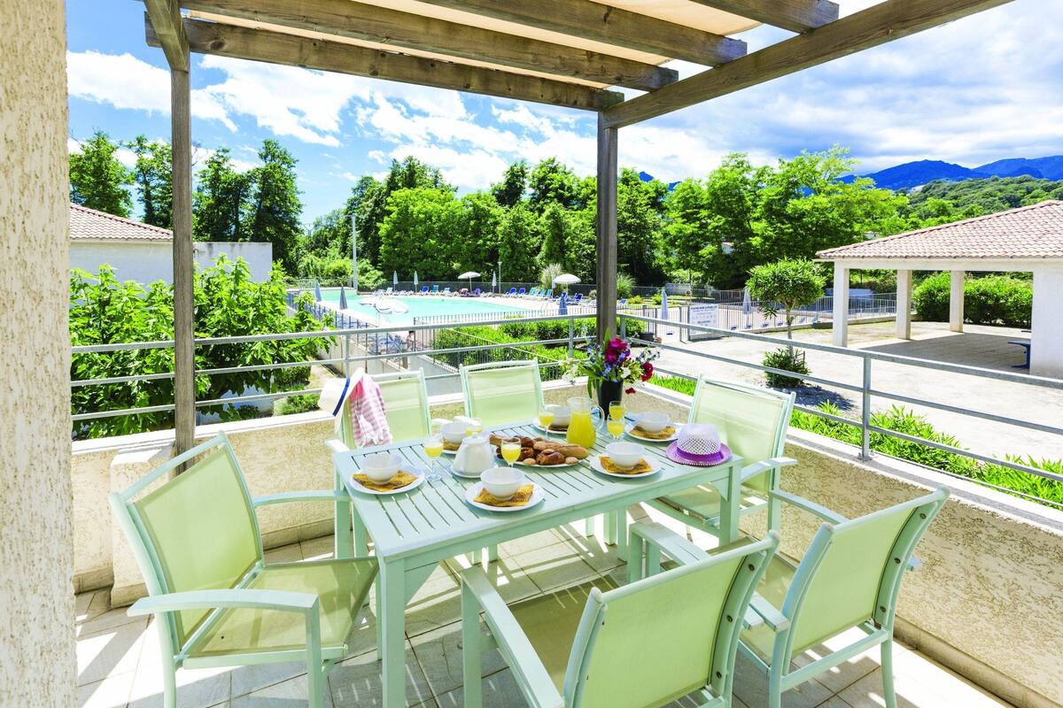 Uma área de refeições coberta ao ar livre, com vista para a piscina, cadeiras verdes e uma mesa posta para o café da manhã.