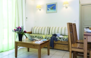 A living room with a sofa, a wooden table with a laptop, a potted plant, and a wall-mounted lamp. - Residence Odalys Acqua Linda (Poggio-Mezzana)