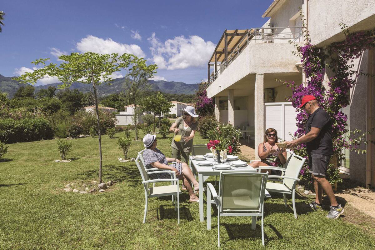 Uma área de refeições ao ar livre com uma mesa posta para quatro pessoas, cercada por vegetação e com montanhas ao fundo.