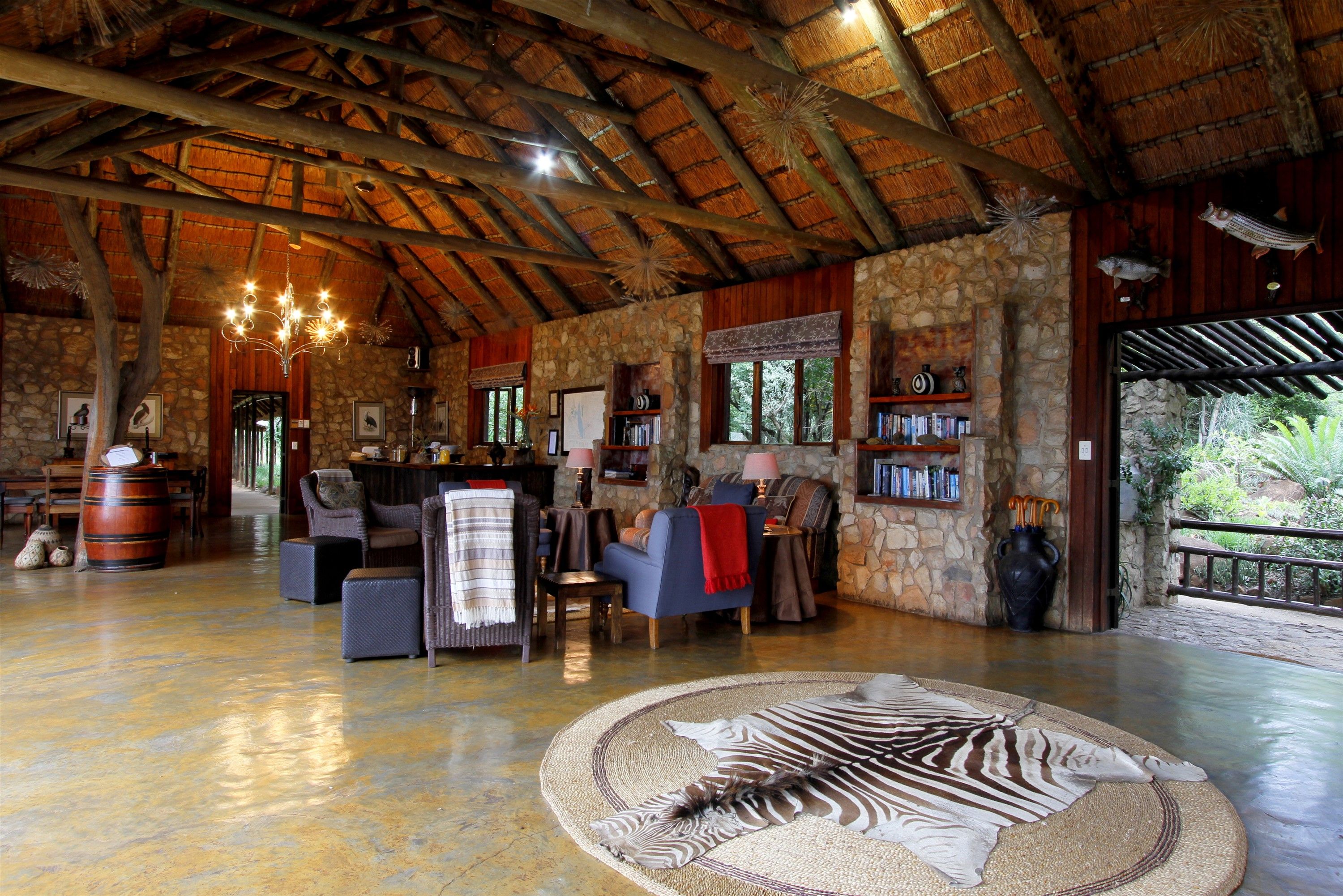 A spacious lounge with a high ceiling, stone walls, and a thatched roof. There are comfortable seating areas, a large rug, and a bookshelf.