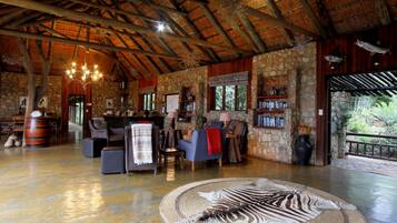 A spacious lounge with a high ceiling, stone walls, and a thatched roof. There are comfortable seating areas, a large rug, and a bookshelf.