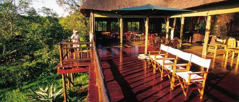 An outdoor dining area with wooden chairs and tables, a thatched roof structure, and a view of the surrounding greenery.