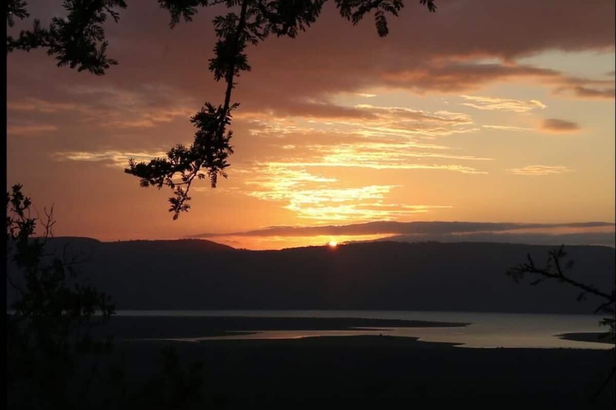 Een zonsondergang boven een wateroppervlak, met silhouetten van bomen en verafgelegen heuvels.