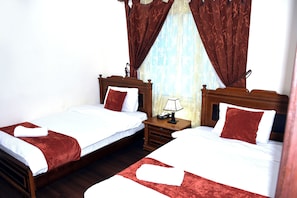 Quarto duplo com duas camas, cabeceiras de madeira, mesa de cabeceira com luminária e janela com cortinas.