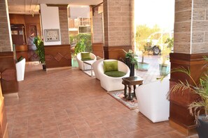 Um hall com piso de azulejos, sofás brancos com almofadas verdes, plantas em vasos e um balcão de recepção.
