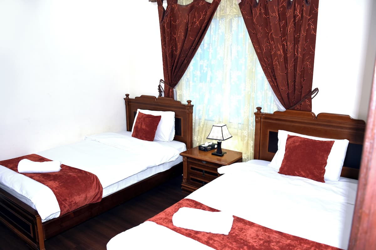 A double room with two beds, wooden headboards, a nightstand with a lamp, and a window with curtains.