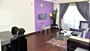 A living room with a purple wall, a sofa, a coffee table, a TV, and a dining area with chairs and a table. - Arinza Tower Quality Suites (Salmiya)