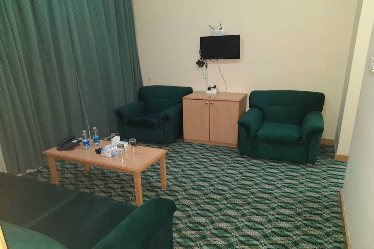 Quarto de hotel com duas poltronas verdes, uma mesa de madeira com telefone e garrafas de água, e uma televisão fixada na parede.