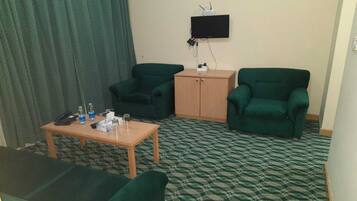 Ein Hotelzimmer mit zwei grünen Sesseln, einem Holztisch mit Telefon und Wasserflaschen sowie einem an der Wand befestigten Fernseher.