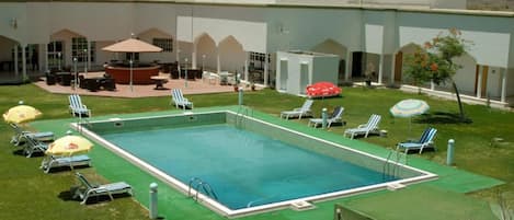Piscina com espreguiçadeiras e guarda-sóis, cercada por um edifício com várias portas e janelas.