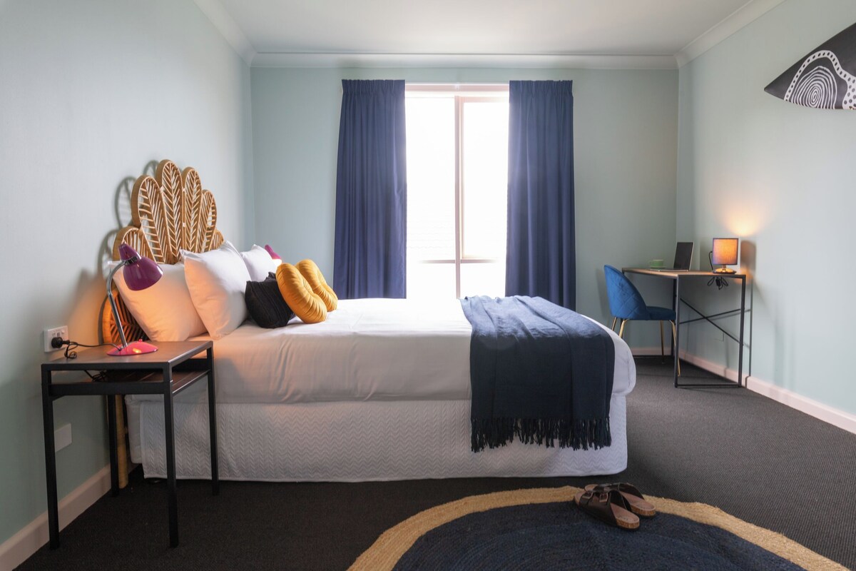 A bedroom with a bed, a desk with a computer, a chair, and a window with curtains.