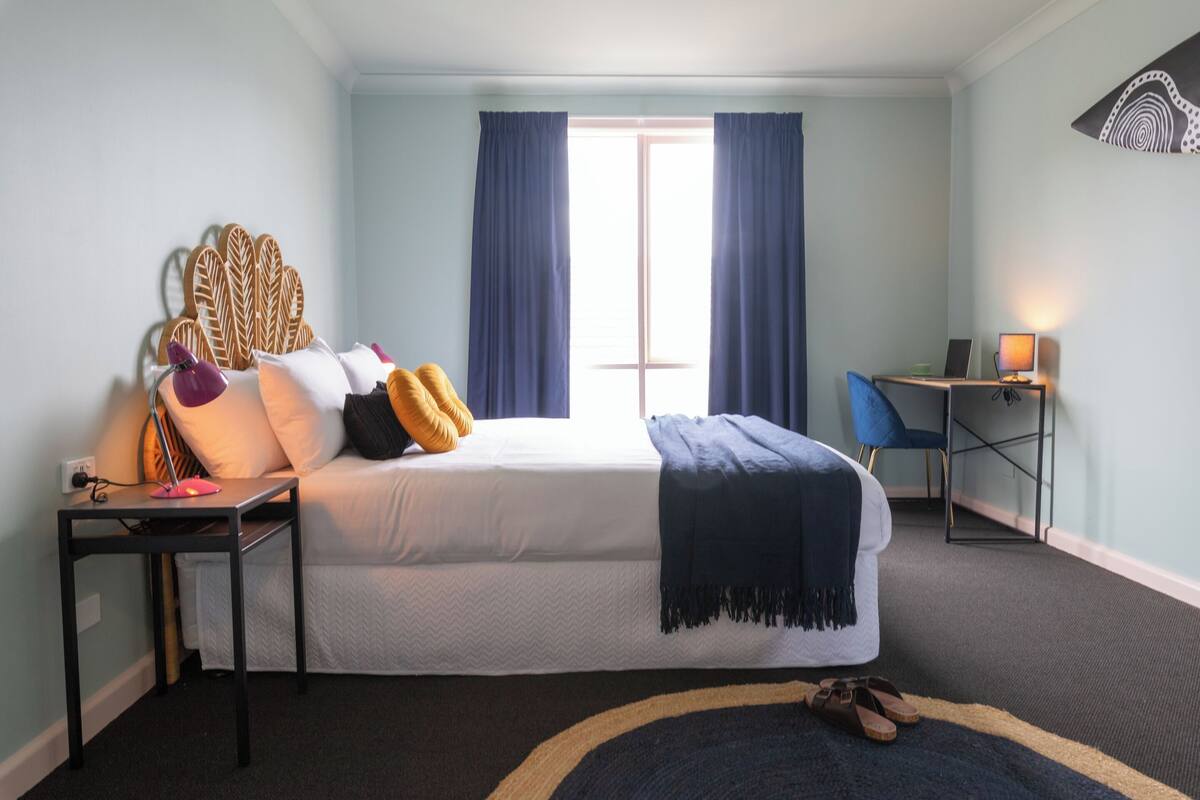 A bedroom with a bed, a desk with a computer, a chair, and a window with curtains.