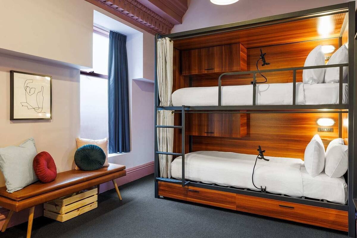 A bunk bed with a wooden headboard, a bench with cushions, a framed picture on the wall, and a window with curtains.