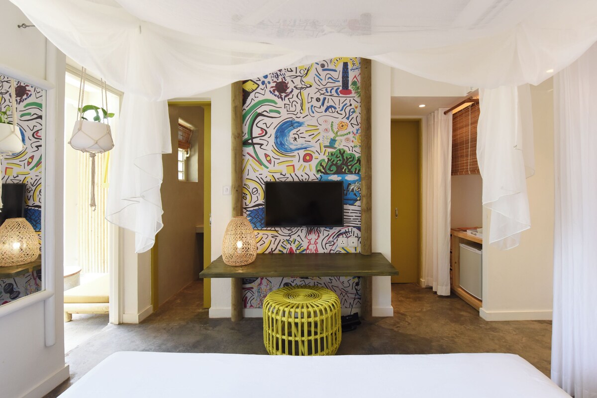Habitación de hotel con un mural abstracto de colores, un televisor de pantalla plana, un taburete tejido y una cama.