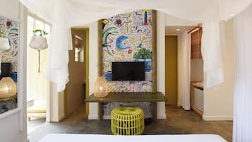 A hotel room with a colorful abstract mural, a flat-screen TV, a woven stool, and a bed.
