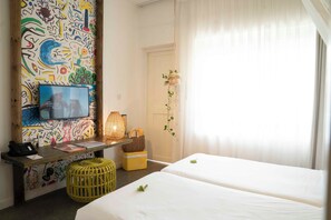 A hotel room with two beds, a desk with a phone and papers, a colorful abstract wall, and a window with sheer curtains. - Veranda Tamarin Hotel & Spa (Tamarin)
