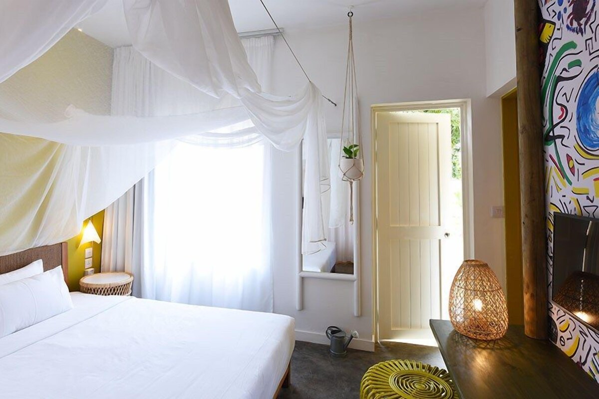 Un dormitorio con cama, una silla colgante, una mesita con una planta y una puerta que da a otra habitación.