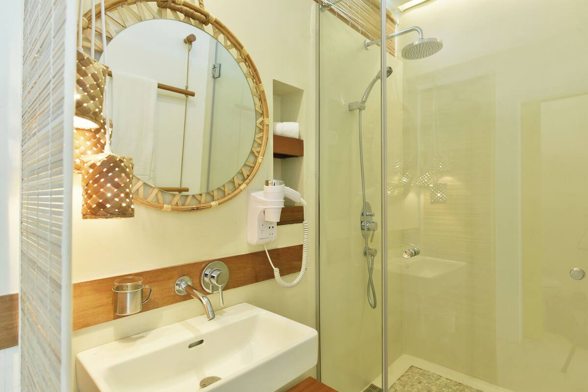 A bathroom with a glass-enclosed shower, a white sink, a round mirror, and a wooden shelf with a towel.