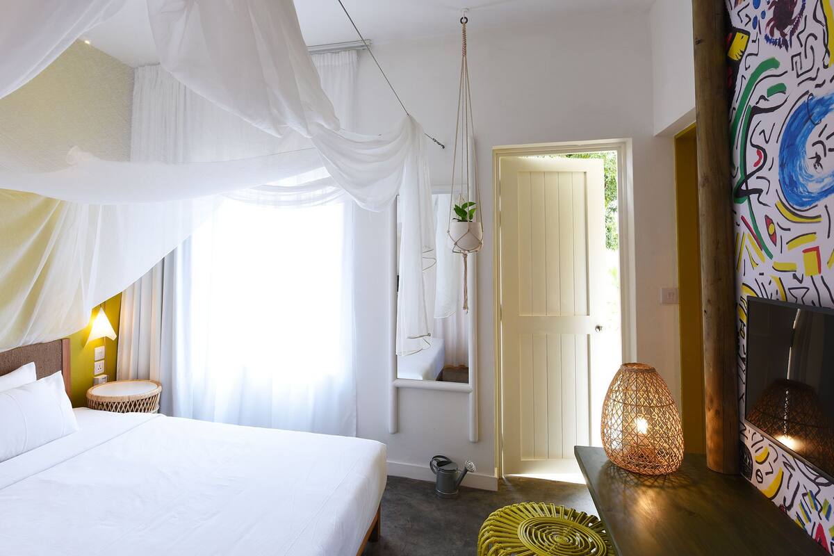 A bedroom with a bed, a bedside lamp, a hanging plant, and a door leading to another room.
