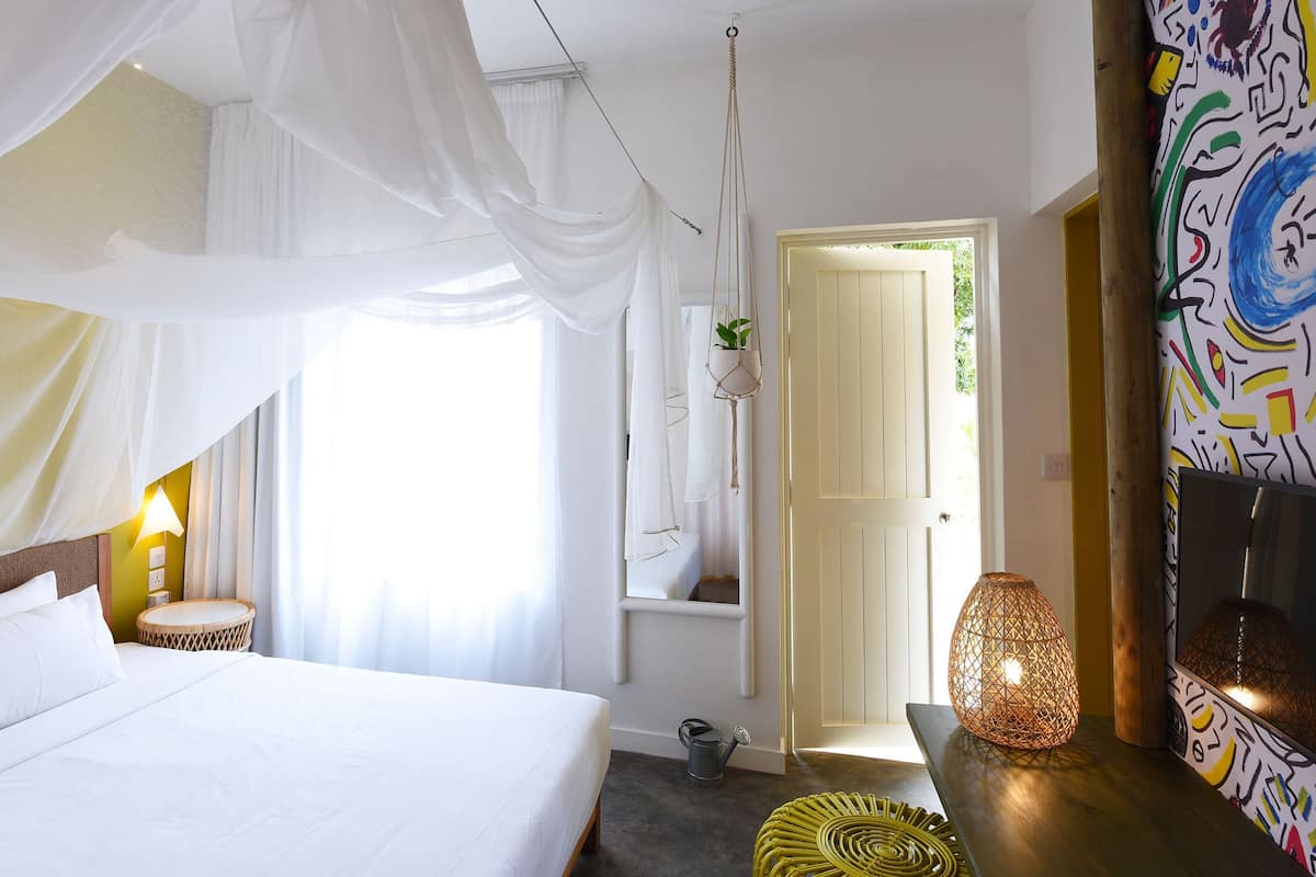 A bedroom with a bed, a bedside lamp, a hanging plant, and a door leading to another room.