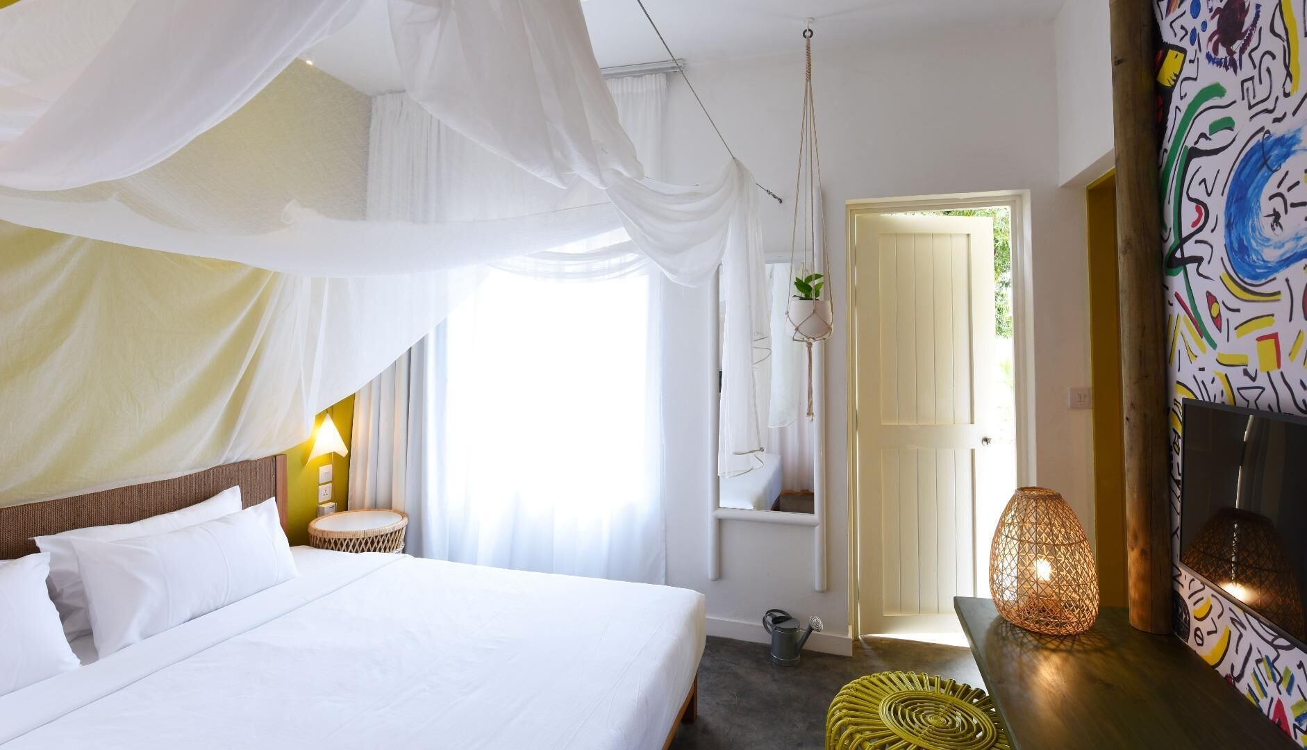 A bedroom with a bed, a bedside lamp, a hanging plant, a door, and a wall with abstract art.