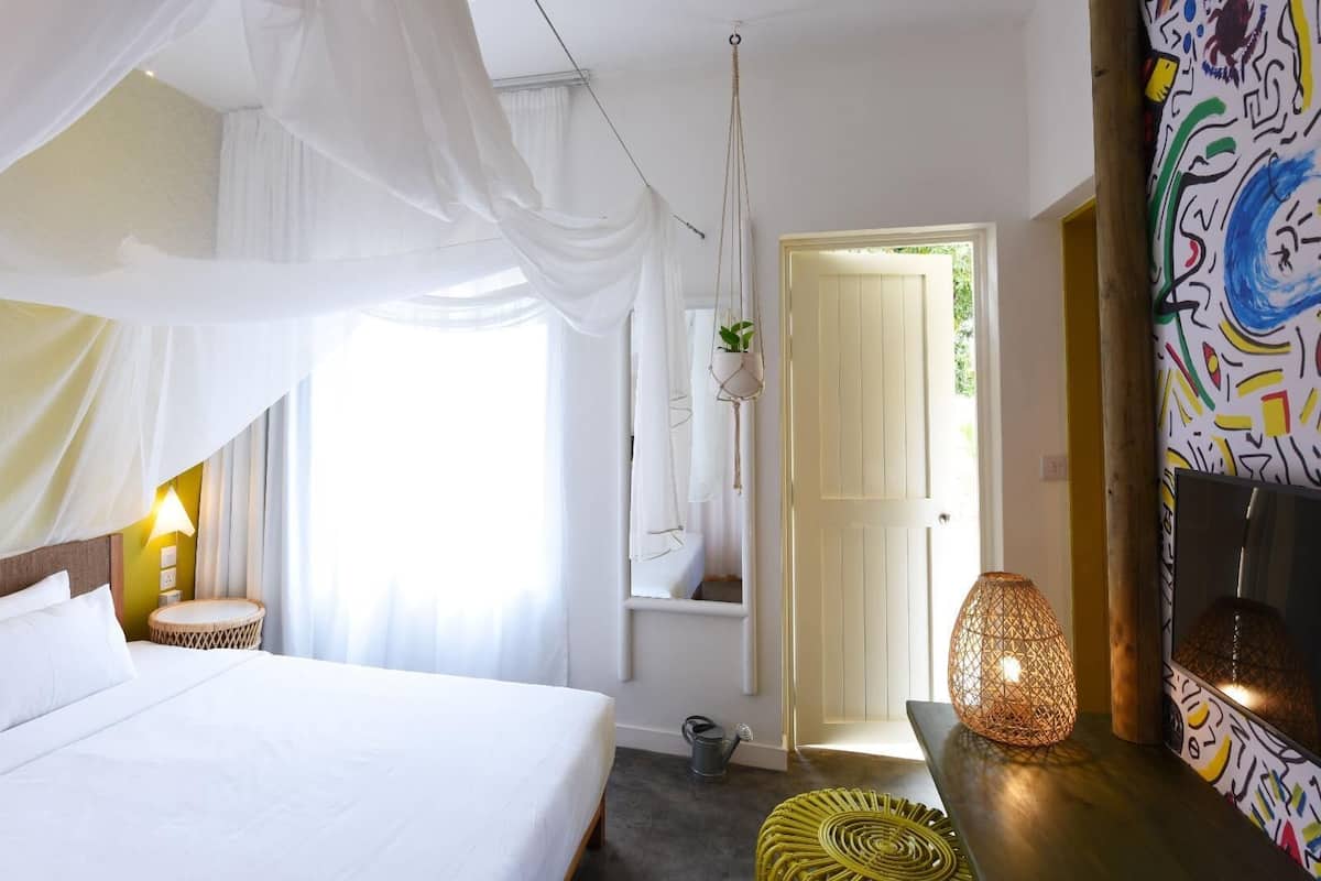 A bedroom with a bed, a bedside lamp, a hanging plant, a door, and a wall with abstract art.