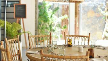 A round dining table with chairs, a hanging light fixture, and a view of greenery outside.