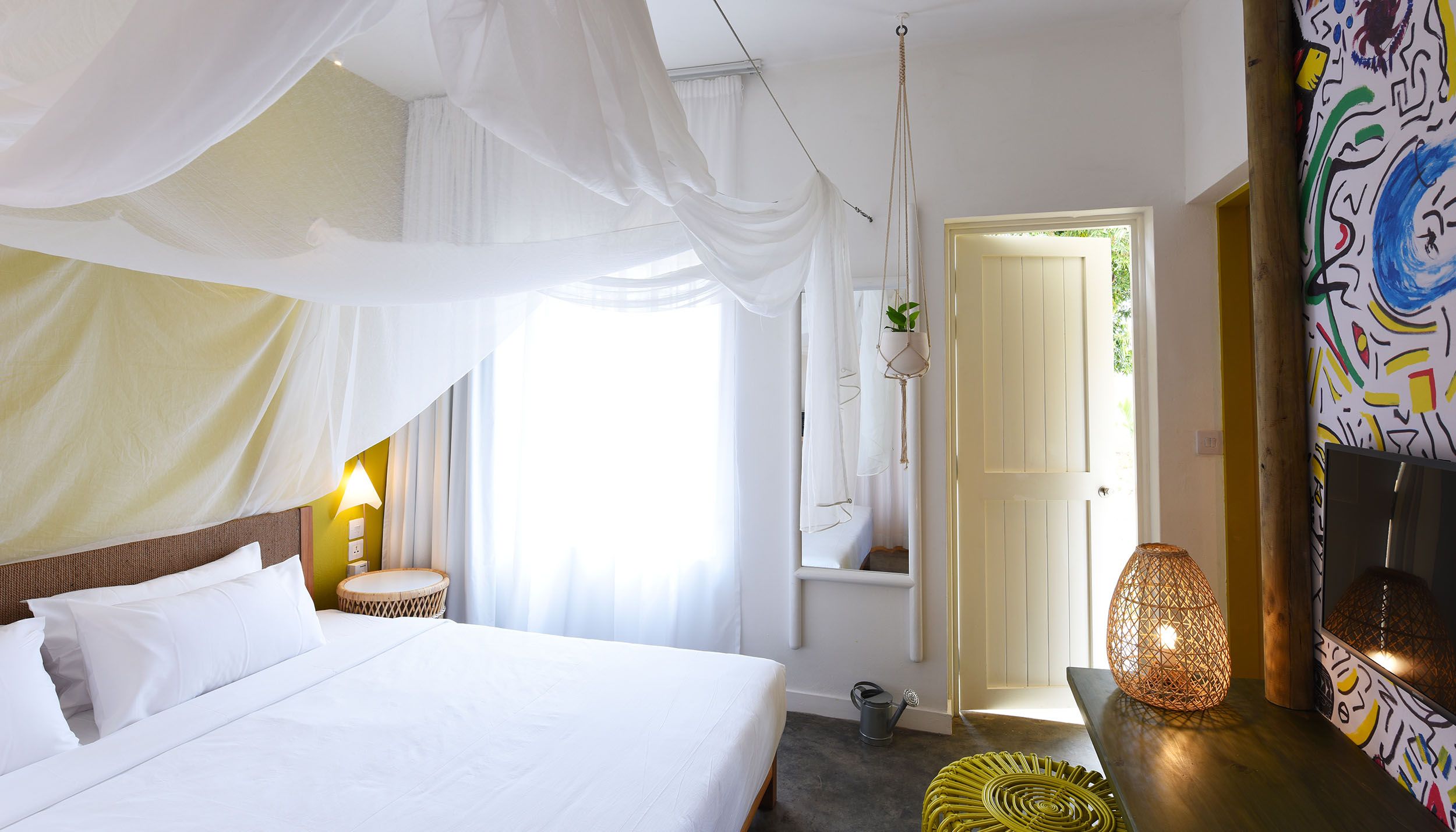 Une chambre à coucher avec un lit, une lampe de chevet, une plante suspendue et une porte donnant sur une autre pièce.