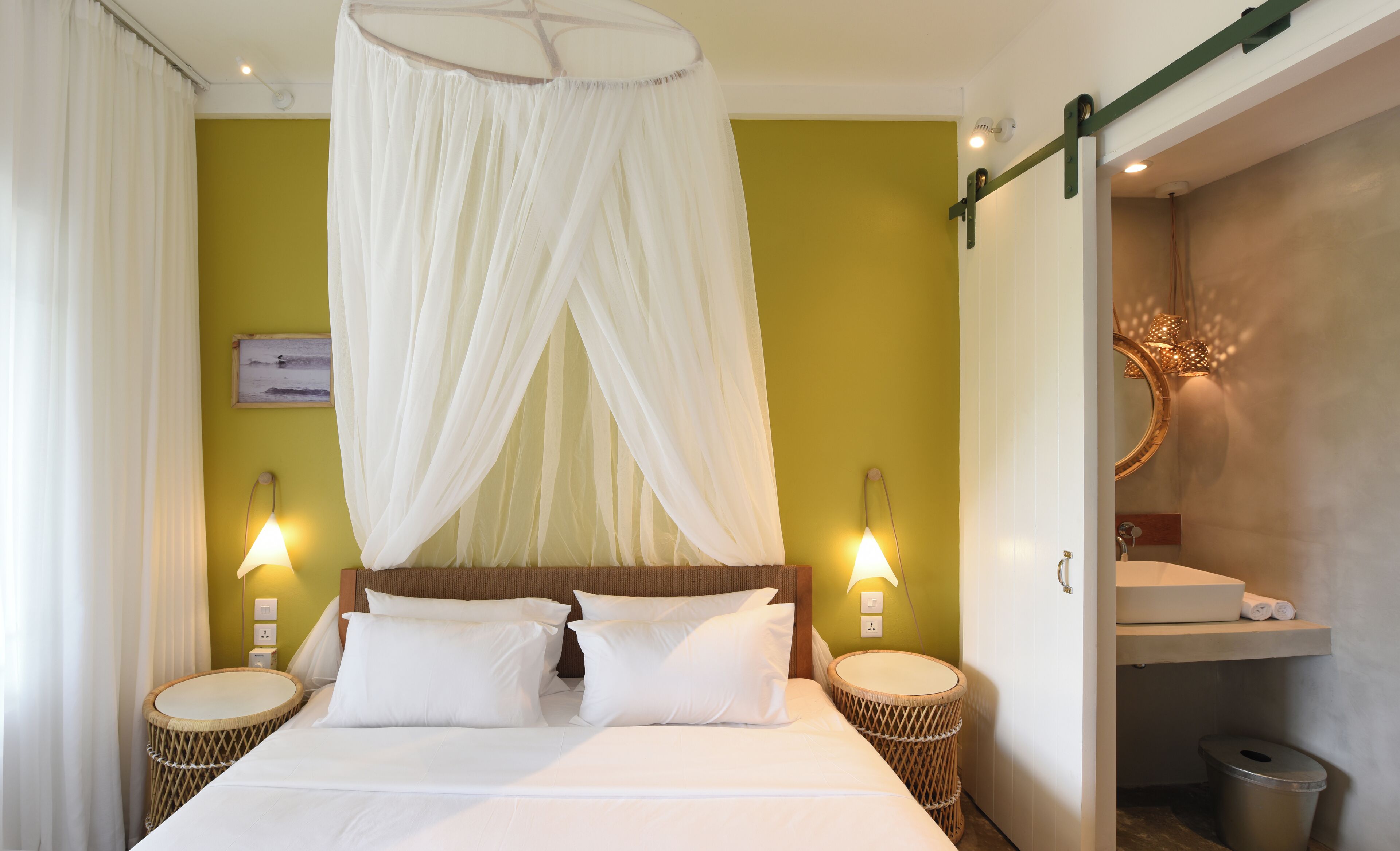 A bed with a canopy, two bedside lamps, a wicker side table, and a wall-mounted mirror.