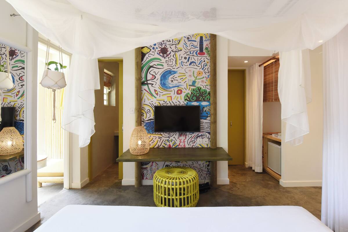 A hotel room with a colorful abstract mural, a flat-screen TV, a woven stool, and a bed.