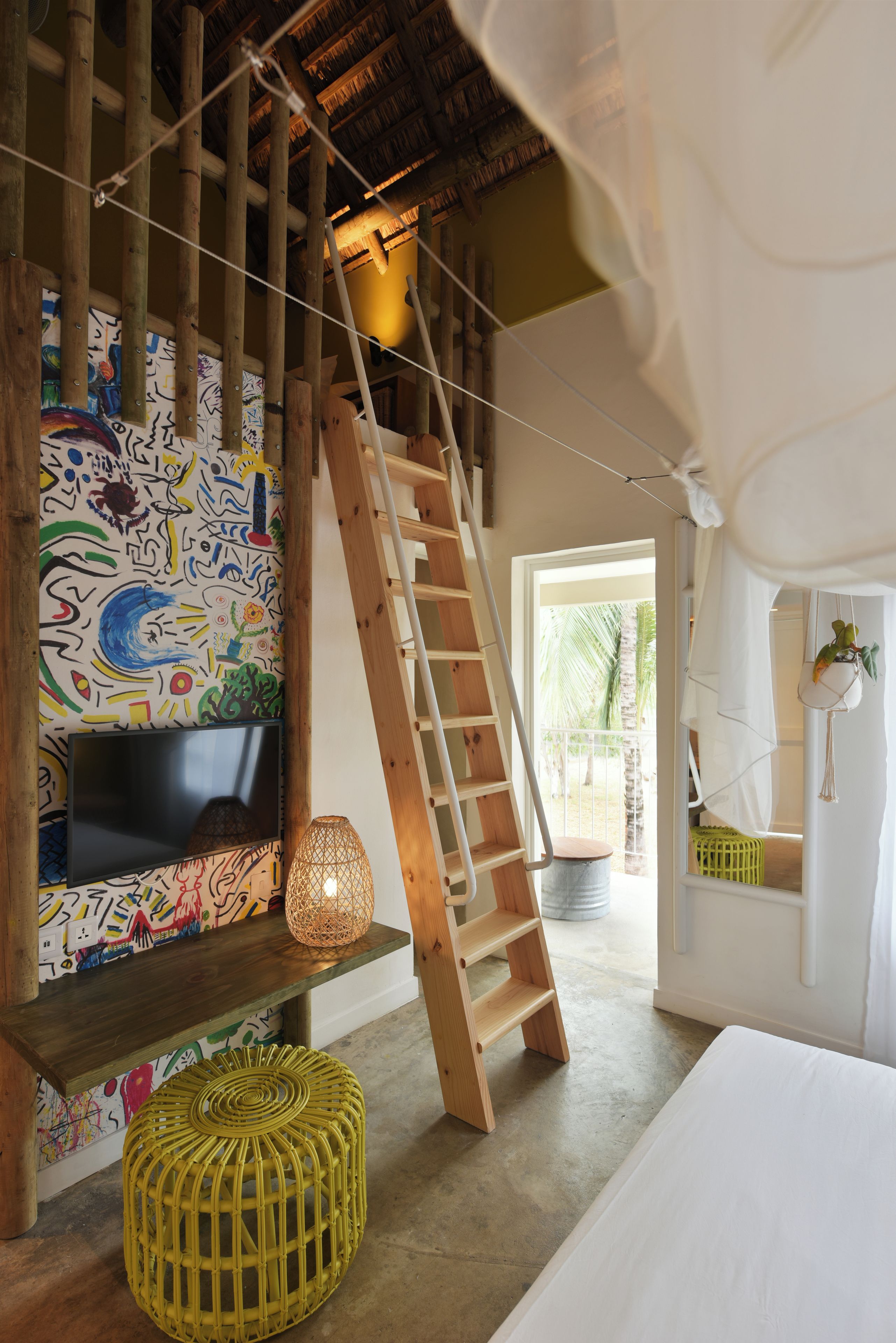 Una habitación con escalera de madera, un mural de pared colorido, una lámpara de pie tejida y una cama con ropa blanca.