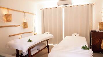 A room with two beds, a wooden bench, a small table, and a wall-mounted air conditioner.