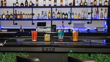 A bar area with a variety of liquor bottles on shelves, a digital screen, and several glasses of mixed drinks on the counter.