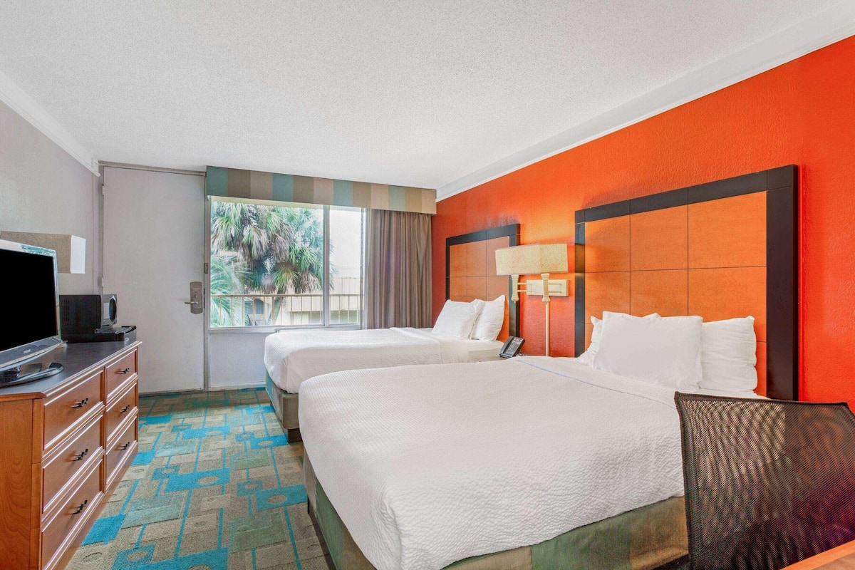 Habitación de hotel con dos camas, televisión, cómoda y ventana con vistas a palmeras.