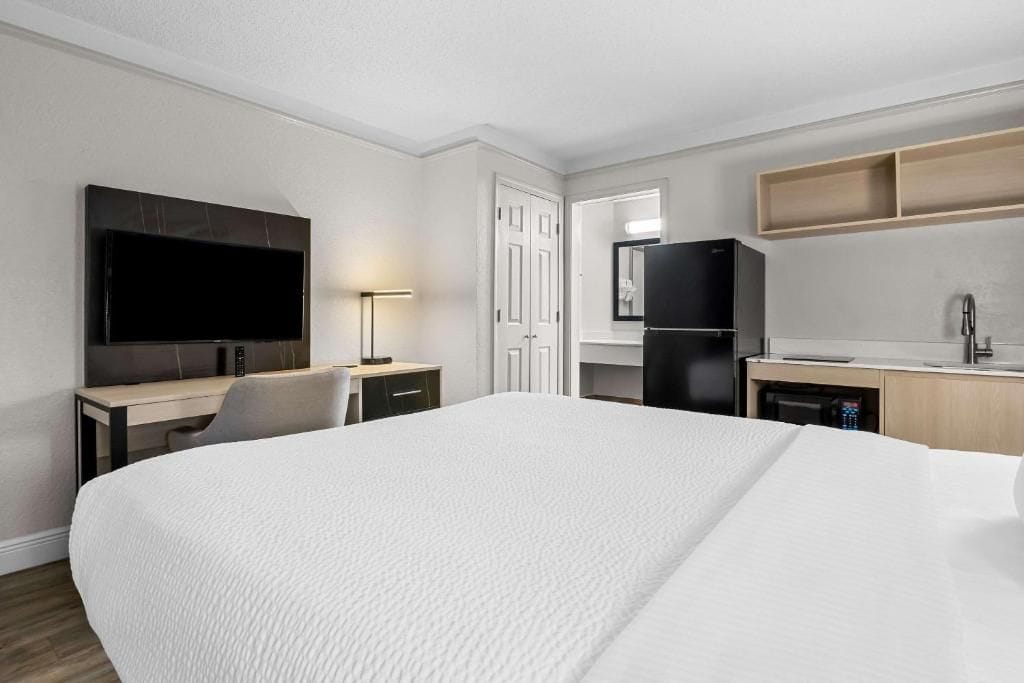 A hotel room with a large bed, a desk with a chair, a television, a refrigerator, a sink, and a mirror.